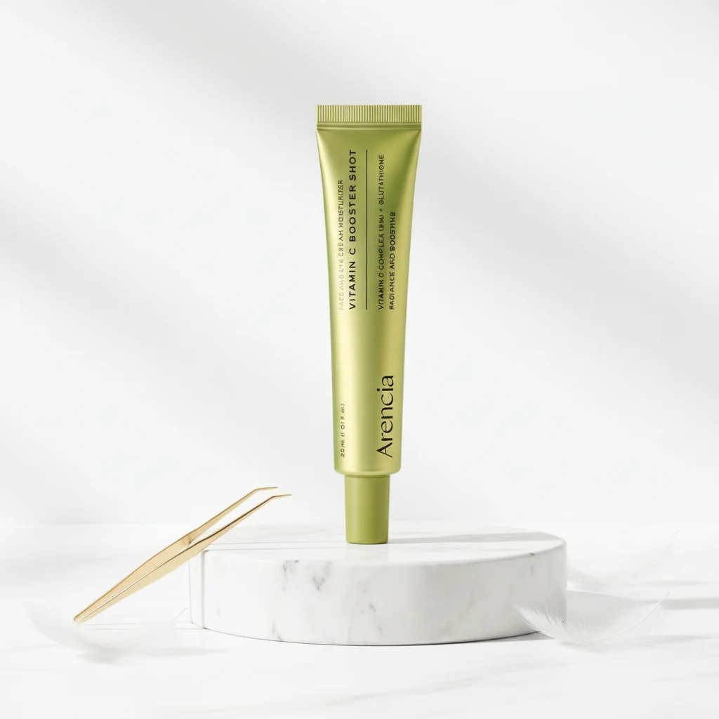 A gold squeeze tube of Arencia Vitamin C Booster Shot stands upright on a white marble pedestal, with a small gold spatula resting to the left, against a clean white background.