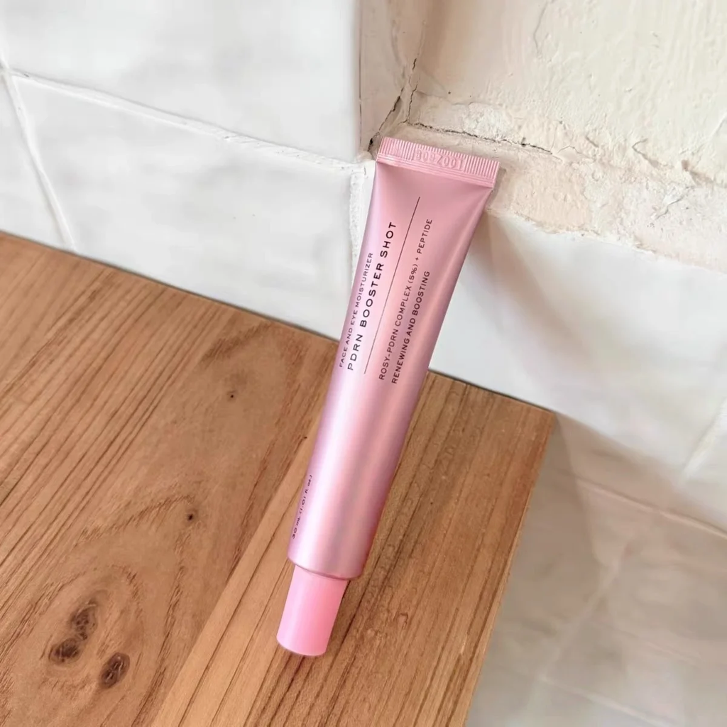 A pink squeeze tube of Power Booster Shop Power Reviver Lip Balm resting diagonally on a wooden surface against a white tiled wall.