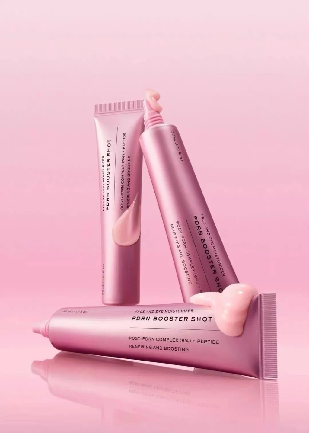 Three pink cosmetic tubes of Rodn Booster Shot skincare product arranged on a reflective pink surface against a solid pink background.