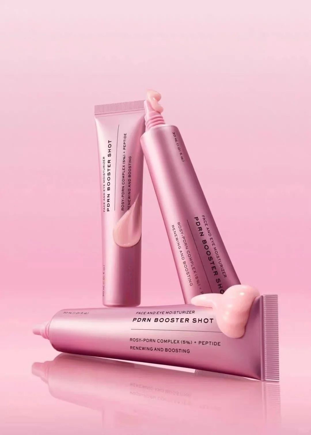 Three pink cosmetic tubes of Rodn Booster Shot skincare product arranged on a reflective pink surface against a solid pink background.