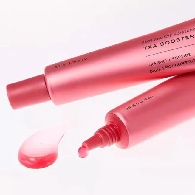 Pink cosmetic tube labeled 'TXA BOOSTER' shown on a white background, with one cap removed and a small droplet of pink product in front.